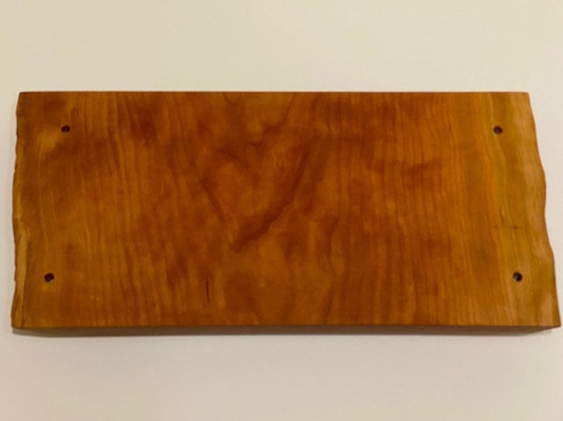 Op de afbeelding: Een rechthoekig houten bord met een natuurlijke houtnerf afwerking. Het bord heeft een licht ruwe, onafgewerkte rand en vier kleine gaten die in het oppervlak zijn geboord.