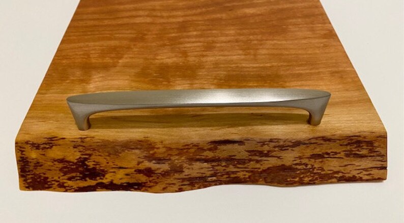Op de afbeelding: Een houten serveerschaal met een natuurlijke houten afwerking en een zilveren metalen handvat. De schaal heeft een natuurlijke rand en is gemaakt van &eacute;&eacute;n stuk hout.