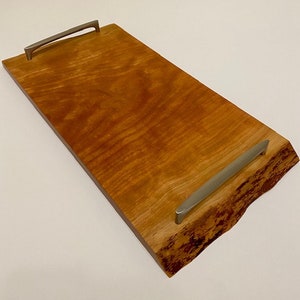 Op de afbeelding: Een rechthoekige houten serveerschaal met een natuurlijke houten afwerking en een levende rand. De schaal heeft twee zilveren metalen handvatten.
