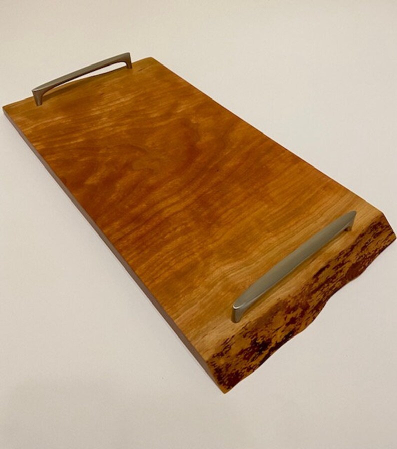 Op de afbeelding: Een rechthoekige houten serveerschaal met een natuurlijke houten afwerking en een levende rand. De schaal heeft twee zilveren metalen handvatten.
