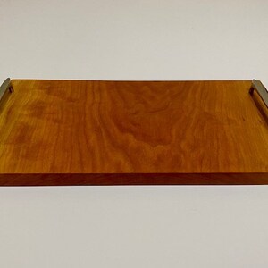 Op de afbeelding: Een rechthoekige houten dienblad met een natuurlijke houten afwerking en twee metalen handvatten. Het dienblad heeft een glad oppervlak en is perfect om eten of drinken te serveren.