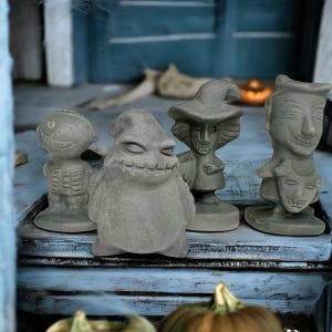 May include: Four gray Halloween figurines with unique character designs. The figures include a skeleton, a character with a stitched mouth, a witch, and a figure with a face and a mask. The figurines are displayed on a blue surface.