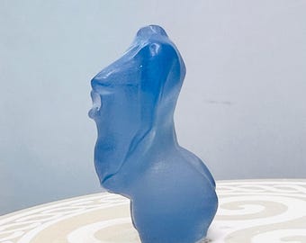 May include: A translucent blue sculpture of a human torso. The sculpture has a smooth, flowing form with subtle details. The piece is displayed on a patterned surface against a light blue background. The sculpture is a unique decorative item.