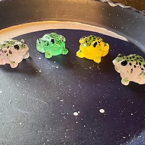 May include: Four small, colorful frog figurines. The frogs are in shades of pink, green, and yellow, with black spots. They are arranged on a dark blue surface with white speckles, resembling a starry night.