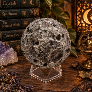 May include: A spherical stone sculpture with a textured, cratered surface in shades of gray, black, and white, resembling the moon. The sphere is supported by a clear, geometric, pyramid-shaped acrylic stand, providing a stable base for display.