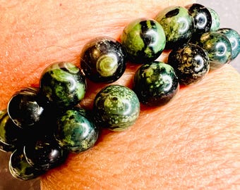 May include: A beaded bracelet featuring round, polished beads in shades of green and black. The beads have a marbled appearance, with swirls and patterns. The bracelet is worn on a wrist, showcasing the jewelry's design and color variations.