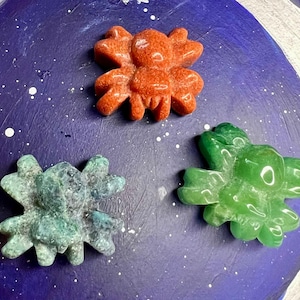 May include: Three carved stone crab figurines in shades of orange, teal, and green rest on a dark blue surface speckled with white. The crab figurines have detailed claw and body features.