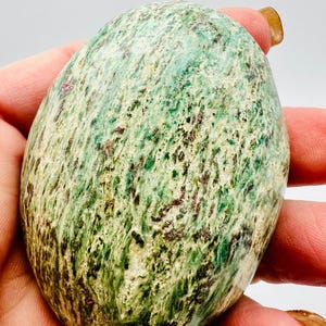May include: A polished, oval-shaped stone with a mottled pattern of green, brown, and cream colors. The stone is held in a hand, showcasing its smooth texture and natural variations. The stone's surface has dark brown and reddish-brown speckles.