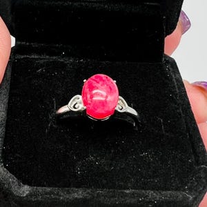 May include: A silver ring with a large, oval-shaped, bright pink gemstone. The ring is set in a simple, elegant design with decorative swirls on either side of the gemstone. The ring is displayed in a black jewelry box.