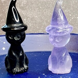 May include: Two carved crystal figurines of cats wearing witch hats. One is black and the other is a translucent purple. The figurines are sitting on a blue surface. The cat figurines have round eyes and are in a sitting position.