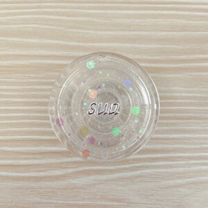 以下が含まれることがあります： 「sua」と銀色の文字が書かれた透明な丸い容器で、内部には虹色のグリッターが入っています。
