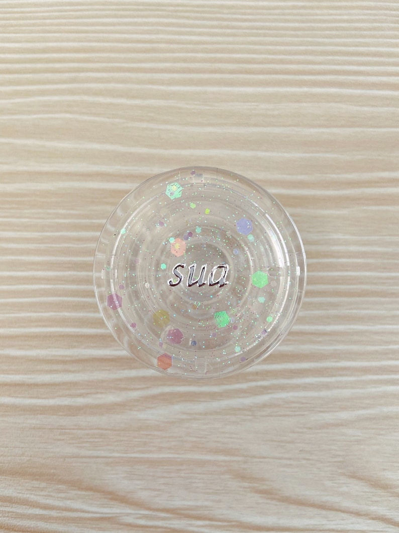 以下が含まれることがあります： 「sua」と銀色の文字が書かれた透明な丸い容器で、内部には虹色のグリッターが入っています。