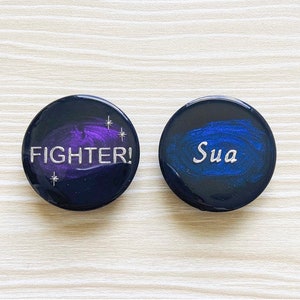 May include: Two black round buttons with a galaxy design. One button has the word "FIGHTER!" in silver lettering. The other button has the word "Sua" in silver lettering.
