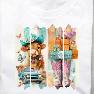 Puede incluir: Una camiseta blanca con un diseño gráfico colorido que presenta una vaca con un sombrero azul y botas, un camión y una valla con mariposas. El diseño es de estilo acuarela.