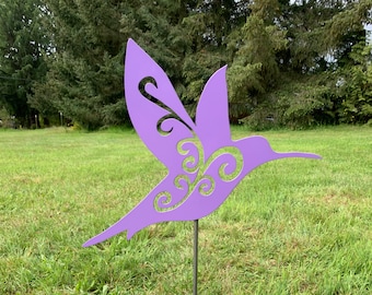 May include: A purple metal hummingbird garden stake with decorative cut-out details. The hummingbird is depicted in flight, mounted on a black metal pole, and set against a backdrop of green grass and trees. A decorative lawn ornament.