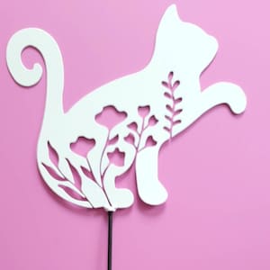 May include: White metal silhouette of a cat with floral cutouts on a pink background. The cat is standing on a black metal stick.