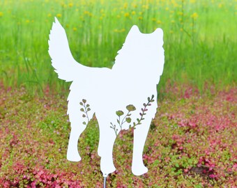May include: A white metal dog silhouette garden stake with floral accents. The dog-shaped decoration is placed in a garden bed with red and green foliage, with a backdrop of green grass and trees.