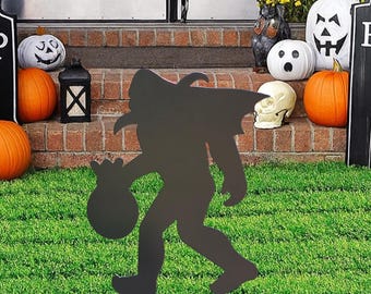 May include: A black metal Bigfoot silhouette garden stake, holding a sack, is positioned in front of a Halloween display. The display features pumpkins, a skull, and a tombstone with the words "There Is Beetlejuice".