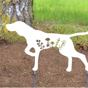 Puede incluir: Una estaca de jardín de metal blanco con forma de silueta de perro con recortes florales. El perro está en posición de caza, con una pata levantada. La estaca está insertada en el suelo, con un árbol y hierba verde en el fondo.