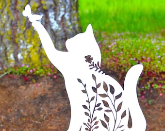 May include: A white metal cat silhouette garden stake with a butterfly on its outstretched paw. The cat's body features a cut-out floral design. The stake is placed in a garden setting with a tree and green grass in the background.