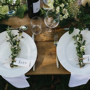 May include: A wedding table setting with white plates, cutlery, and floral decorations. Each plate features a bundle of herbs and a name tag tied with string. A wine bottle and glasses are also visible.