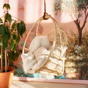 A Sedona Moonrise hanging rattan chair hangs in a well-lit room with pink wallpaper and a potted plant in the corner. The hanging rattan chair has a white cushion and white pillows and a white fur blanket added for decoration.