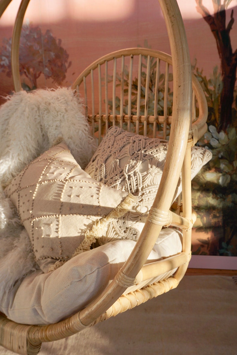 A Sedona Moonrise hanging rattan chair hangs in a well-lit room with pink wallpaper and a potted plant in the corner. The hanging rattan chair has a white cushion and white pillows and a white fur blanket added for decoration.