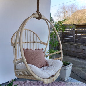 A light-colored rattan hanging chair on a patio with potted plants. The Sedona Moonrise hanging chair has a white cushion and a pink pillow with pom-poms on the seat for decoration with a pink rug below.