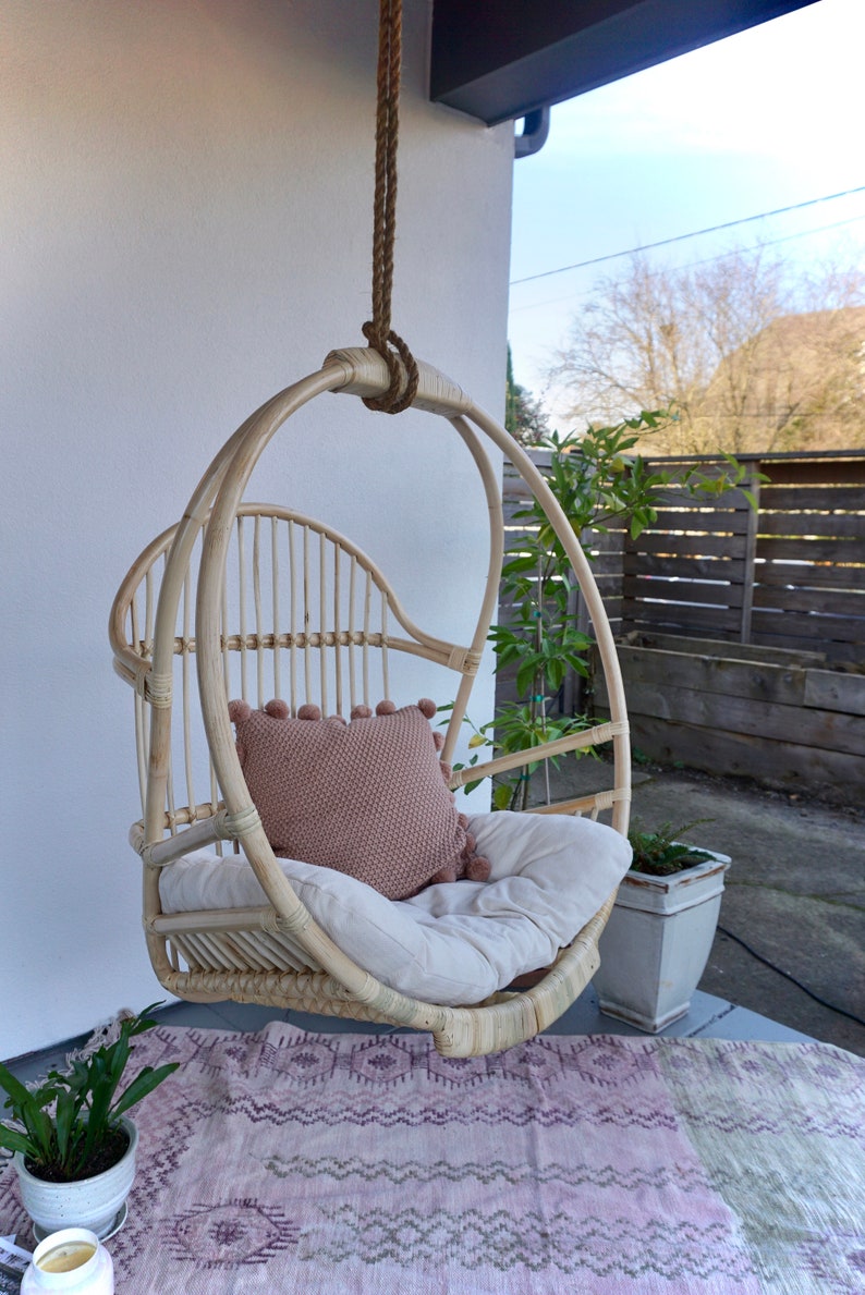 A light-colored rattan hanging chair on a patio with potted plants. The Sedona Moonrise hanging chair has a white cushion and a pink pillow with pom-poms on the seat for decoration with a pink rug below.