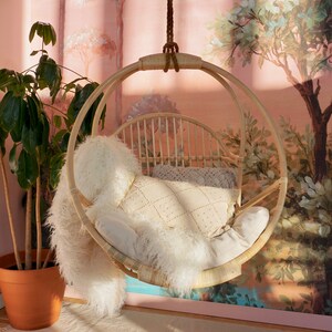 A Sedona Moonrise hanging rattan chair hangs in a well-lit room with pink wallpaper and a potted plant in the corner. The hanging rattan chair has a white cushion and white pillows and a white fur blanket added for decoration.