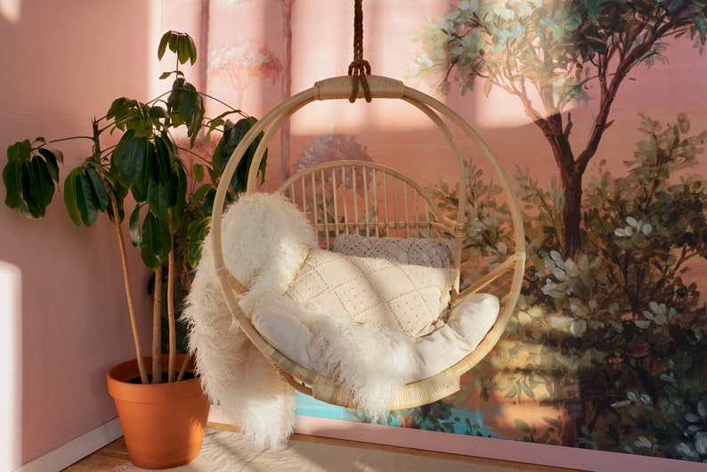 A Sedona Moonrise hanging rattan chair hangs in a well-lit room with pink wallpaper and a potted plant in the corner. The hanging rattan chair has a white cushion and white pillows and a white fur blanket added for decoration.