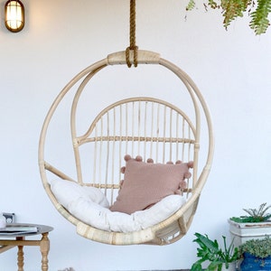 A light-colored rattan hanging chair on a patio with potted plants and a side table with reading materials. The Sedona Moonrise hanging chair has a white cushion and a pink pillow with pom-poms on the seat for decoration.