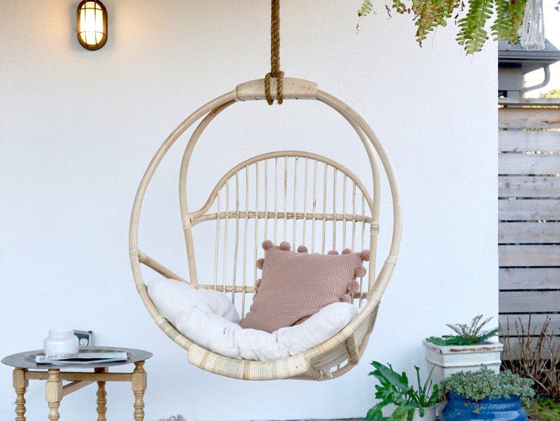 A light-colored rattan hanging chair on a patio with potted plants and a side table with reading materials. The Sedona Moonrise hanging chair has a white cushion and a pink pillow with pom-poms on the seat for decoration.