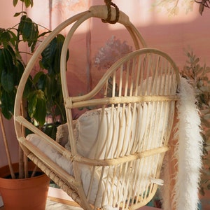 A Sedona Moonrise hanging rattan chair hangs in a well-lit room with pink wallpaper and a potted plant in the corner. The back of the chair has multiple supports for leisurely sitting and highlights a white fur blanket added for decoration.