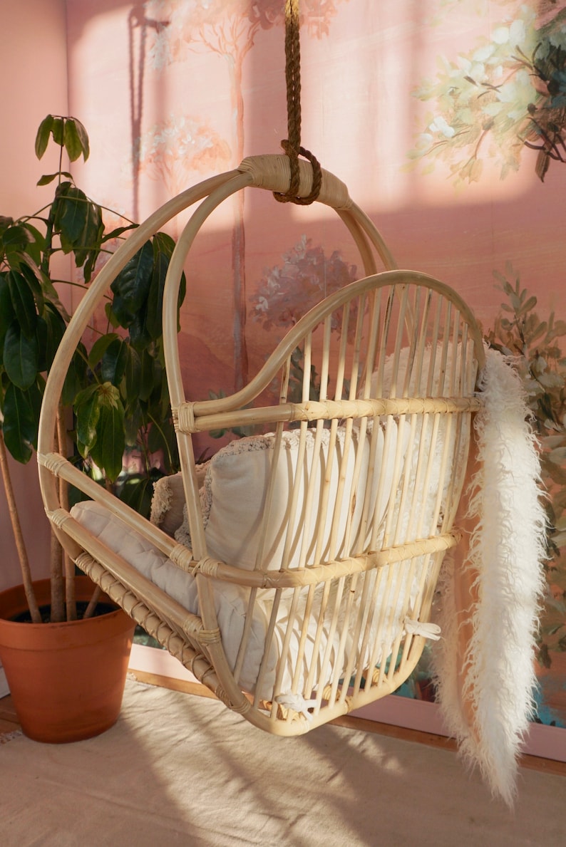 A Sedona Moonrise hanging rattan chair hangs in a well-lit room with pink wallpaper and a potted plant in the corner. The back of the chair has multiple supports for leisurely sitting and highlights a white fur blanket added for decoration.