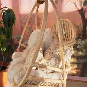 A Sedona Moonrise hanging rattan chair hangs in a well-lit room with pink wallpaper and a potted plant in the corner. The hanging rattan chair has a white cushion and white pillows and a white fur blanket added for decoration.