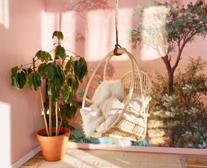 A Sedona Moonrise hanging rattan chair hangs in a well-lit room with pink wallpaper and a potted plant in the corner. The hanging rattan chair has a white cushion and white pillows and a white fur blanket added for decoration.