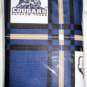 May include: A folded blue, black, and beige plaid blanket. The blanket features a white square with the text "COUGARS BRIGHAM YOUNG" and a cougar logo. The blanket is in a clear plastic bag.