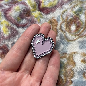 May include: A pink pixelated heart-shaped enamel pin with a black outline.