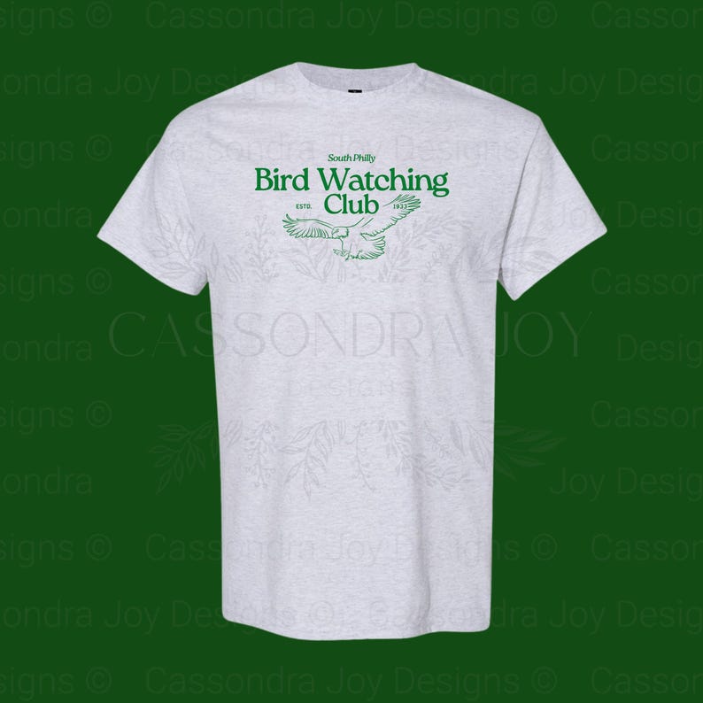 Puede incluir: Una camiseta gris jaspeado claro con un gr&aacute;fico verde en el pecho. El texto dice South Philly Bird Watching Club ESTD 1922, acompa&ntilde;ado de una ilustraci&oacute;n lineal de un p&aacute;jaro en vuelo con las alas extendidas ampliamente.