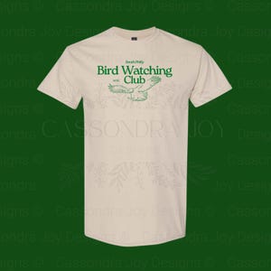 Puede incluir: Una camiseta de manga corta de color beige con texto verde que dice South Philly Bird Watching Club EST. 1973, acompa&ntilde;ada de una ilustraci&oacute;n gr&aacute;fica verde de un p&aacute;jaro en vuelo centrada en la zona del pecho de la prenda.