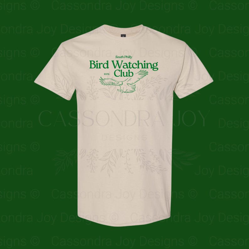 Puede incluir: Una camiseta de manga corta de color beige con texto verde que dice South Philly Bird Watching Club EST. 1973, acompa&ntilde;ada de una ilustraci&oacute;n gr&aacute;fica verde de un p&aacute;jaro en vuelo centrada en la zona del pecho de la prenda.