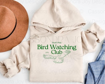 Puede incluir: Una sudadera con capucha de color beige que presenta el texto verde South Philly Bird Watching Club ESTD. 1923 y un gráfico de un águila volando. La sudadera se muestra junto a un sombrero de fieltro marrón y vaqueros azules.