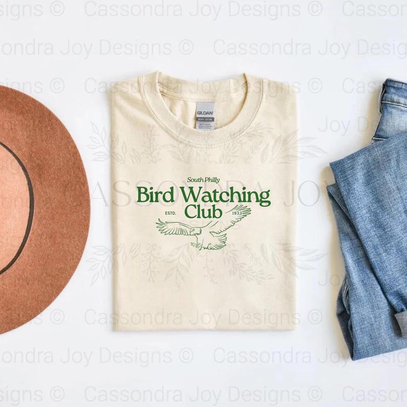 Puede incluir: Una camiseta de manga corta color crema con un estampado gr&aacute;fico verde que dice South Philly Bird Watching Club ESTD 1923 junto a una ilustraci&oacute;n lineal de un p&aacute;jaro en vuelo. La camiseta est&aacute; colocada junto a un sombrero marr&oacute;n y unos vaqueros azules.