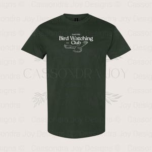 Puede incluir: Una camiseta de manga corta de color verde bosque oscuro con texto blanco que dice South Philly Bird Watching Club ESTD 1922, acompa&ntilde;ada de una ilustraci&oacute;n de l&iacute;neas blancas de un p&aacute;jaro con las alas extendidas en el centro del pecho.