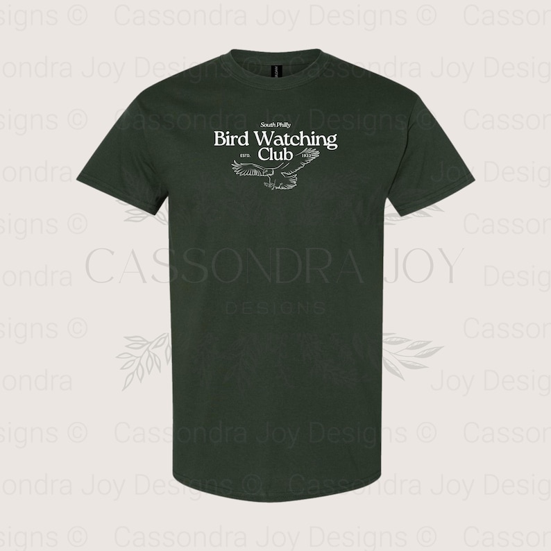 Puede incluir: Una camiseta de manga corta de color verde bosque oscuro con texto blanco que dice South Philly Bird Watching Club ESTD 1922, acompa&ntilde;ada de una ilustraci&oacute;n de l&iacute;neas blancas de un p&aacute;jaro con las alas extendidas en el centro del pecho.