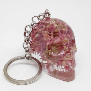 May include: A clear resin skull keychain with pink and white pressed flowers inside. The keychain has a silver metal ring and chain.