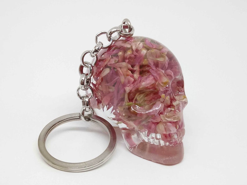 May include: A clear resin skull keychain with pink and white pressed flowers inside. The keychain has a silver metal ring and chain.