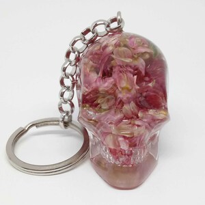 May include: A clear resin skull keychain with pink flower petals inside. The keychain has a silver metal ring and chain.