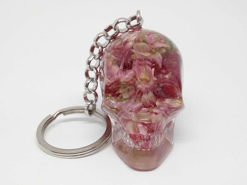 May include: A clear resin skull keychain with pink flower petals inside. The keychain has a silver metal ring and chain.